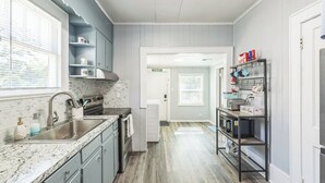 A modern kitchen with a granite countertop, stainless steel sink, and a built-in oven. There is a window with blinds, a door leading to another room, and a shelf with kitchen utensils. - Cozy Bungalow near Downtown Pensacola: Game Room, Patio + Fire Pit, 3 Bedroom! (Pensacola)