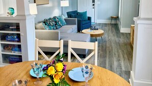 A dining area with a wooden table set for two, a sofa with teal cushions, and a ceiling fan. - Cozy Bungalow near Downtown Pensacola: Game Room, Patio + Fire Pit, 3 Bedroom! (Pensacola)