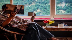 A person reading a book in a wicker chair by a window with a view of a forest. - AUREVELLE KASTURA RESORT KANATAL (Kanatal)
