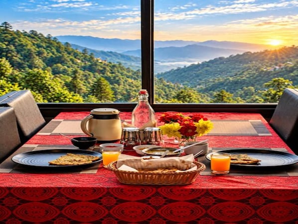 A breakfast setup with a table, plates, a basket, a bottle, and a view of mountains.