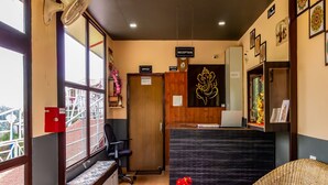 A reception area with a reception desk, a wooden bench, a wicker sofa, and a small round table with flowers. - AUREVELLE KASTURA RESORT KANATAL (Kanatal)
