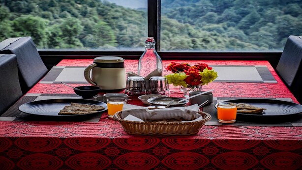 A dining table set with plates, glasses, a bottle, and a basket of bread, with a mountain view through a window. - AUREVELLE KASTURA RESORT KANATAL (Kanatal)