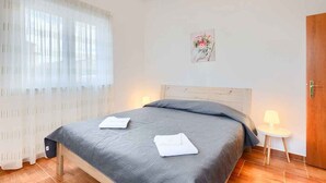 A neatly made bed with a grey bedspread and two white towels, a small bedside table with a lamp, and a wooden door.