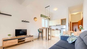 A modern living room with a flat-screen TV, wooden entertainment unit, grey sofa, and a dining area with a table and chairs.