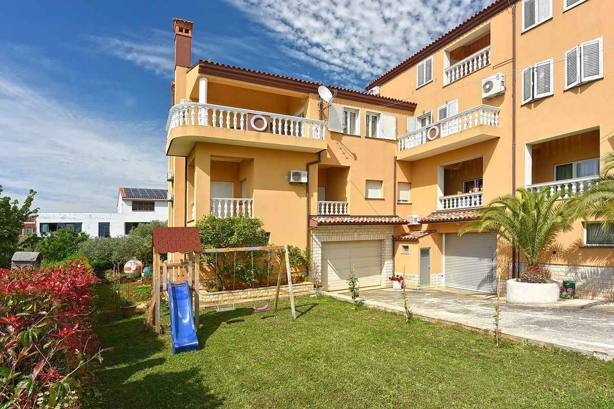 Un edificio residenziale a più piani con balconi, un'area giochi per bambini con uno scivolo e un giardino curato.