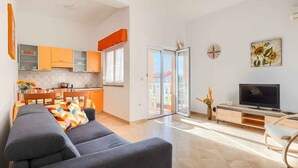 Een moderne woonruimte met een grijze bank, oranje keukenkastjes, een televisie en een glazen schuifdeur naar een balkon.