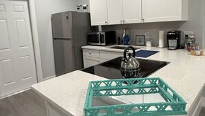 A modern kitchen with white cabinetry, a stainless steel refrigerator, a microwave, and a kettle on the countertop. - Turtle Cove | Steps from the Beach 2BR w/Hockey/Ping-pong/Pool Table (Cocoa Beach)