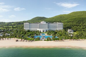 A large, multi-story hotel with a swimming pool and beach view, surrounded by lush greenery and hills.