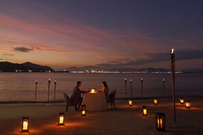 A couple dining at a table with candles on a beach at sunset.