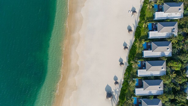 An aerial view of a beachfront resort with multiple bungalow-style buildings, a sandy beach, and clear turquoise waters.