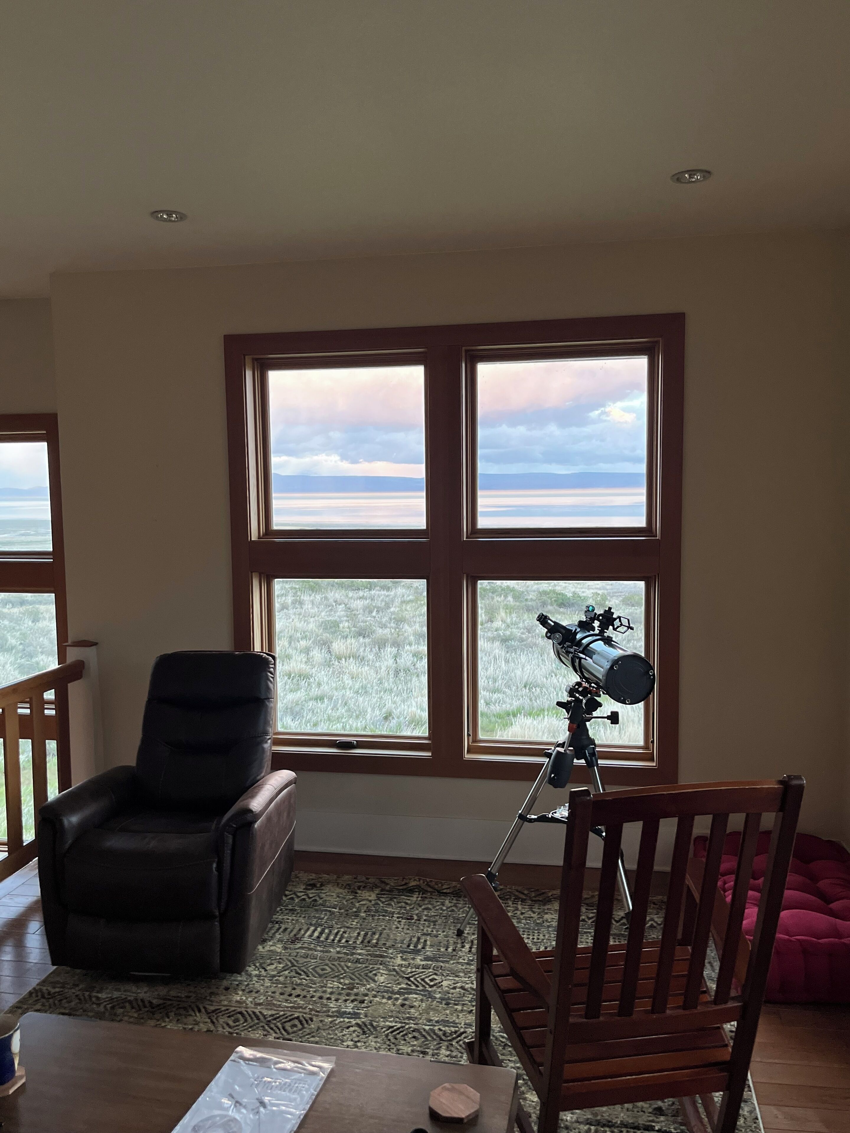 Una habitación con ventanas amplias, un telescopio, un sillón de cuero y una silla de madera.