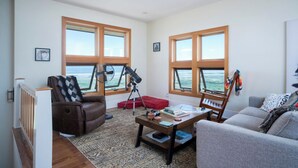 Ein Wohnzimmer mit braunem Ledersofa, einem grauen Sofa, einem Holztisch und einem roten Hocker. Es gibt zwei Fenster mit Holzrahmen, ein Teleskop und einen Ventilator.