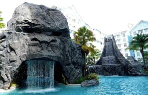 Een zwembad van een resort met een waterval, omringd door grote rotsformaties en palmbomen.