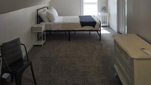 Un dormitorio con cama, una silla, tocador, mesita de noche y ventana.