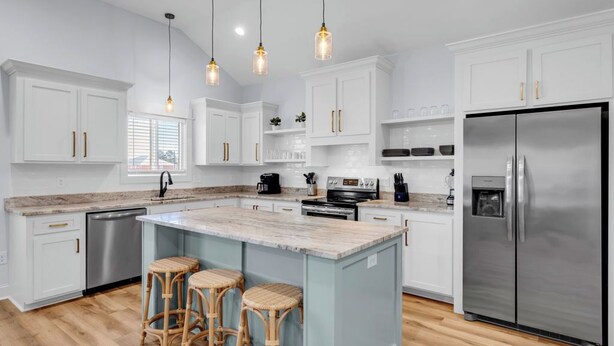 A modern kitchen with white cabinetry, a central island with stools, stainless steel appliances, and pendant lighting. - Waterfront Escape in Bay STL with dock,pickleball, ping pong, basketball & more! (Bay St. Louis)