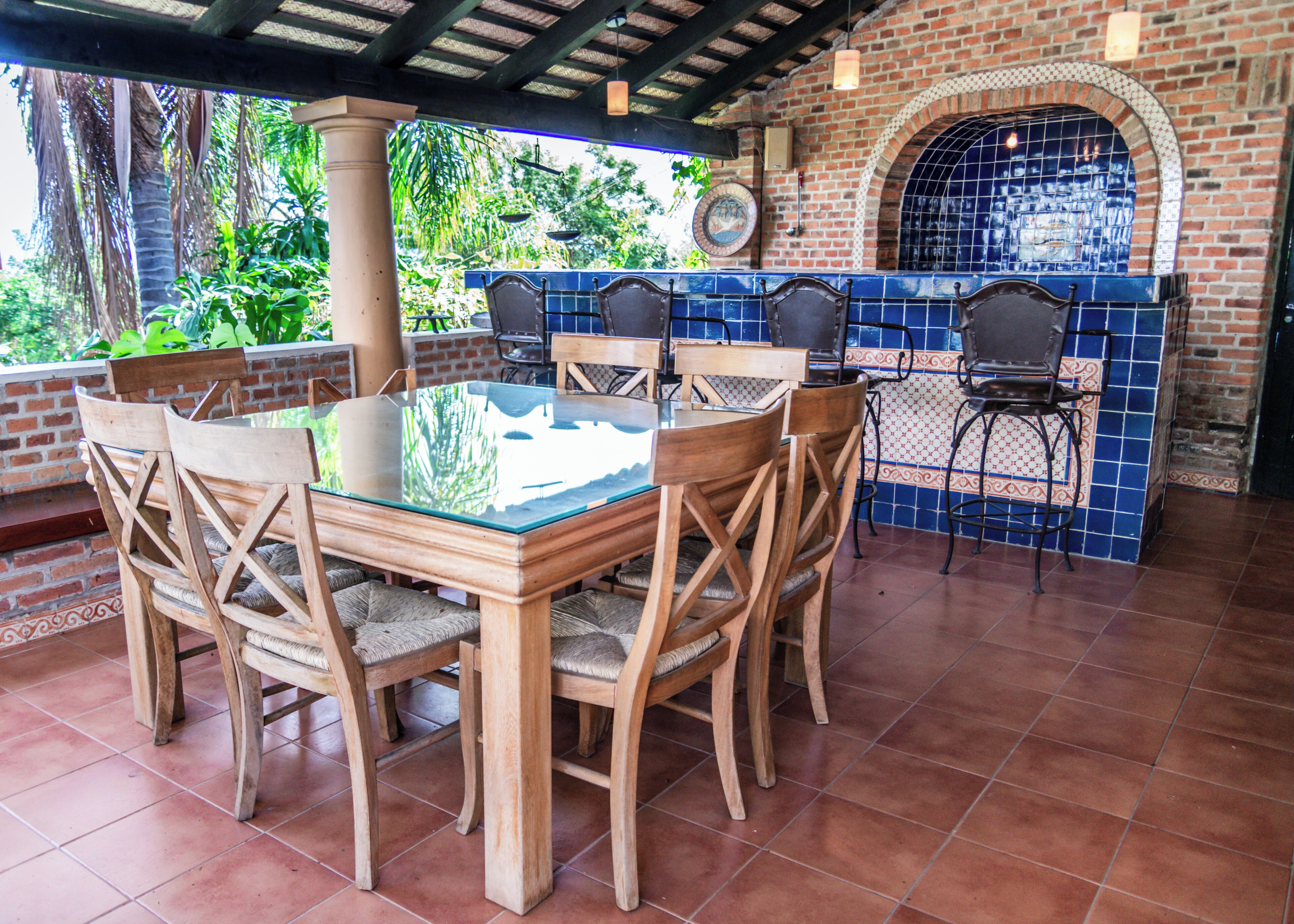 An outdoor patio with a glass-top table surrounded by wooden chairs, a brick wall with a tiled arch, and a view of greenery.