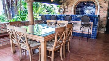 An outdoor patio with a glass-top table surrounded by wooden chairs, a brick wall with a tiled arch, and a view of greenery.