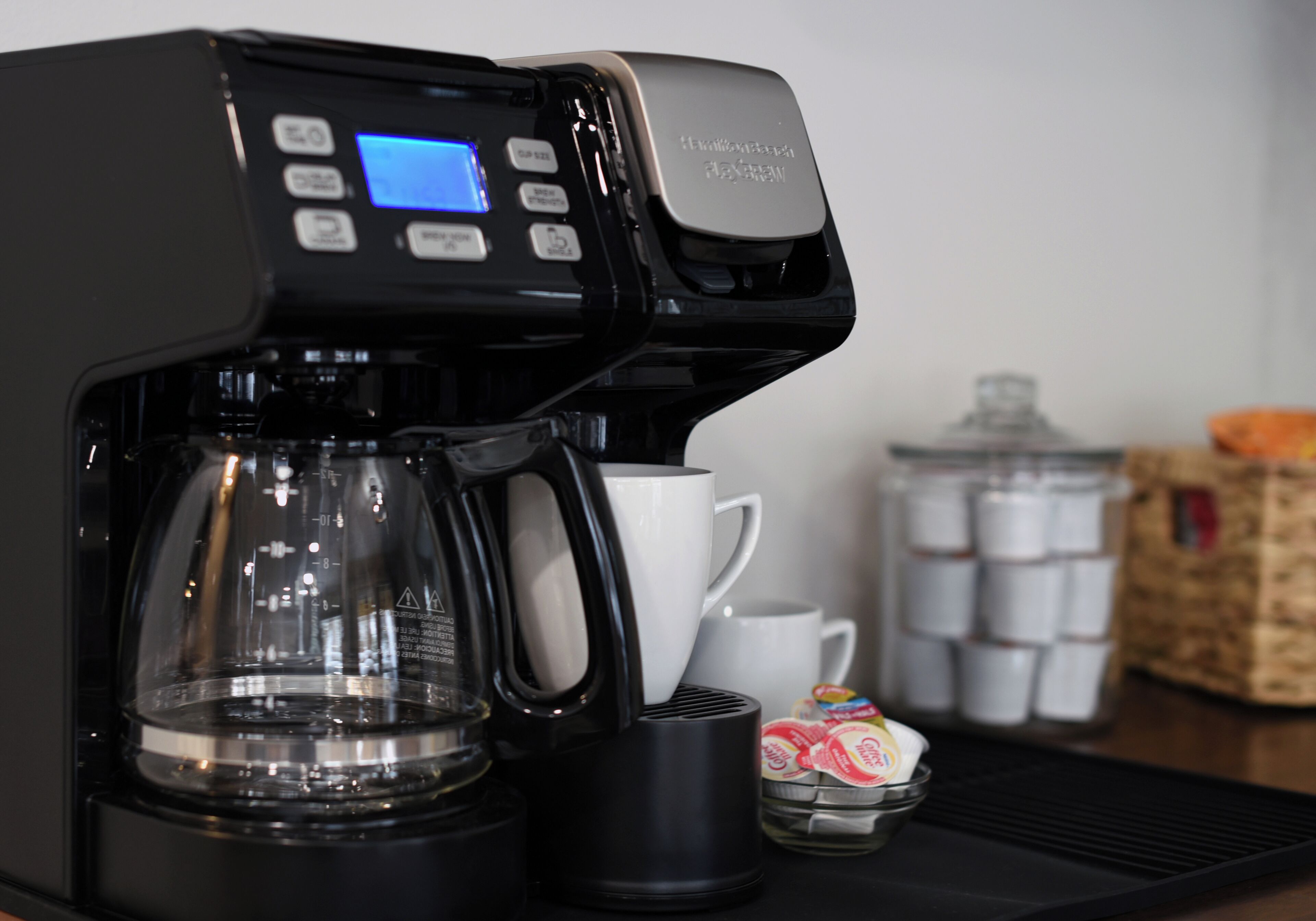 Une machine à café dotée d’un écran numérique, d’une verseuse en verre, d’une tasse blanche et d’un récipient contenant des capsules de café.
