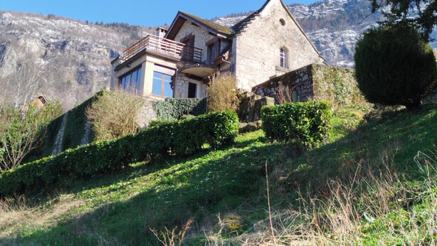 Una casa de pedra amb un balcó, envoltada de verdura i muntanyes al fons.