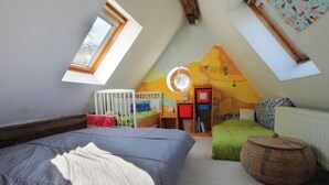 Habitació infantil amb sostre inclinat, una bressol, un llit, una taula petita i una cistella rodona de vimet.