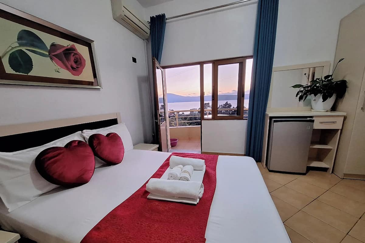 A hotel room with a bed, a desk, a refrigerator, a potted plant, and a view of the sea.