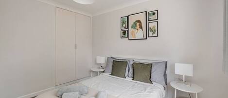 A neatly arranged bedroom with a bed, pillows, a bedside table, and a wall-mounted lamp.