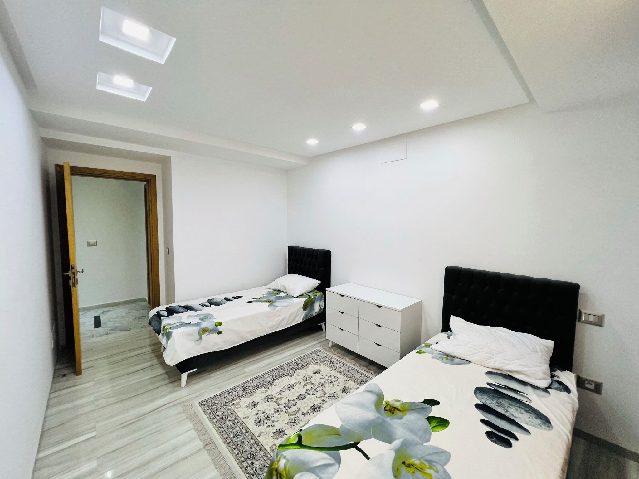 Quarto moderno com duas camas, cômoda branca e tapete estampado.