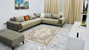 A modern living room with a sectional sofa, a matching ottoman, a patterned rug, a painting, a ceiling light fixture, and a window with curtains. - Tranquil Mrezga Getaway: Near Beach & Hammamet Fun (Mrezga)