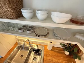 Una cocina con un lavamanos, un escurreplatos y varios cuencos y platos blancos en estantes.