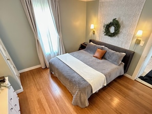 Ein Schlafzimmer mit einem Bett, einem Nachttisch, einer Wandleuchte, einem Fenster mit Vorhängen und einem dekorativen Kranz.