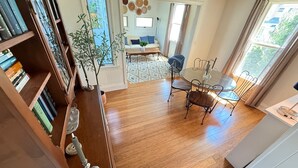 Una habitación con suelo de madera, una mesa de comedor con sillas, una estantería y un ventanal con cortinas.