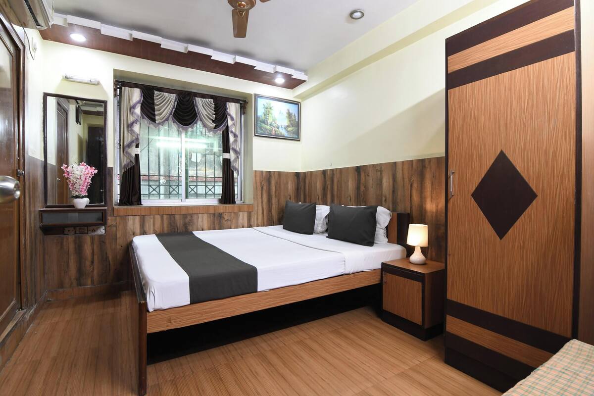 Une chambre à coucher avec un lit en bois, une table de chevet avec une lampe, une fenêtre avec des rideaux, un miroir et une porte ornée d’un motif géométrique.