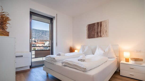 A modern bedroom with a bed, bedside tables, a nightstand, a lamp, and a view of buildings through a window. - 730A Apartment Lumos (Merano)