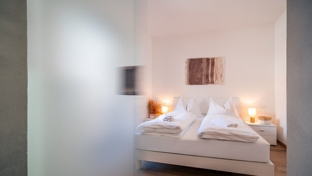 A minimalist bedroom with a bed, bedside tables, and a wall-mounted light fixture. - 730A Apartment Lumos (Merano)