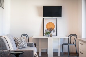 A modern living room with a sofa, two chairs, a white table, a framed artwork, and a flat-screen TV mounted on the wall.