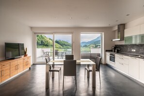 A modern kitchen with a large window, a dining area with a table and chairs, and a television on a wooden cabinet. - 733B Apartment Villa Laurin (Caldaro sulla strada del vino)