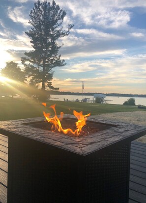 A fire pit with flames, surrounded by a wooden deck, overlooking a body of water with a flag and distant shoreline. - Newer build 3-bedroom cottage on peaceful Fremont Lake with AC, WiFi (Fremont)