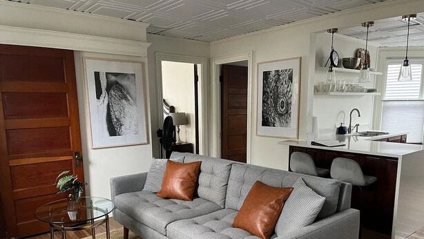 Una sala de estar moderna con un sofá gris, una mesita de vidrio y un tapete tejido. Hay una zona de cocina con encimera blanca e iluminación colgante.