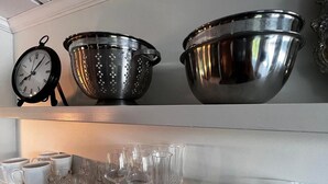 Una repisa de cocina con un colador, un tazón metálico grande y varios vasos, incluyendo copas de vino y tazas.