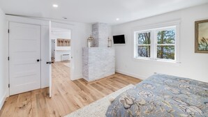 A bedroom with a bed, a white door, a fireplace, a TV, and a window with a view of trees.