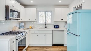 A modern kitchen with white cabinetry, a blue refrigerator, a stove, and a window with a view of trees.