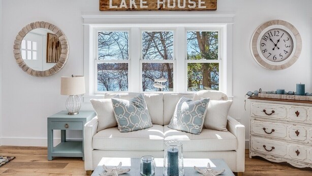 A living room with a white sofa, patterned cushions, a side table with a lamp, a dresser with drawers, a wall clock, and a sign above the windows reading "LAKE HOUSE".