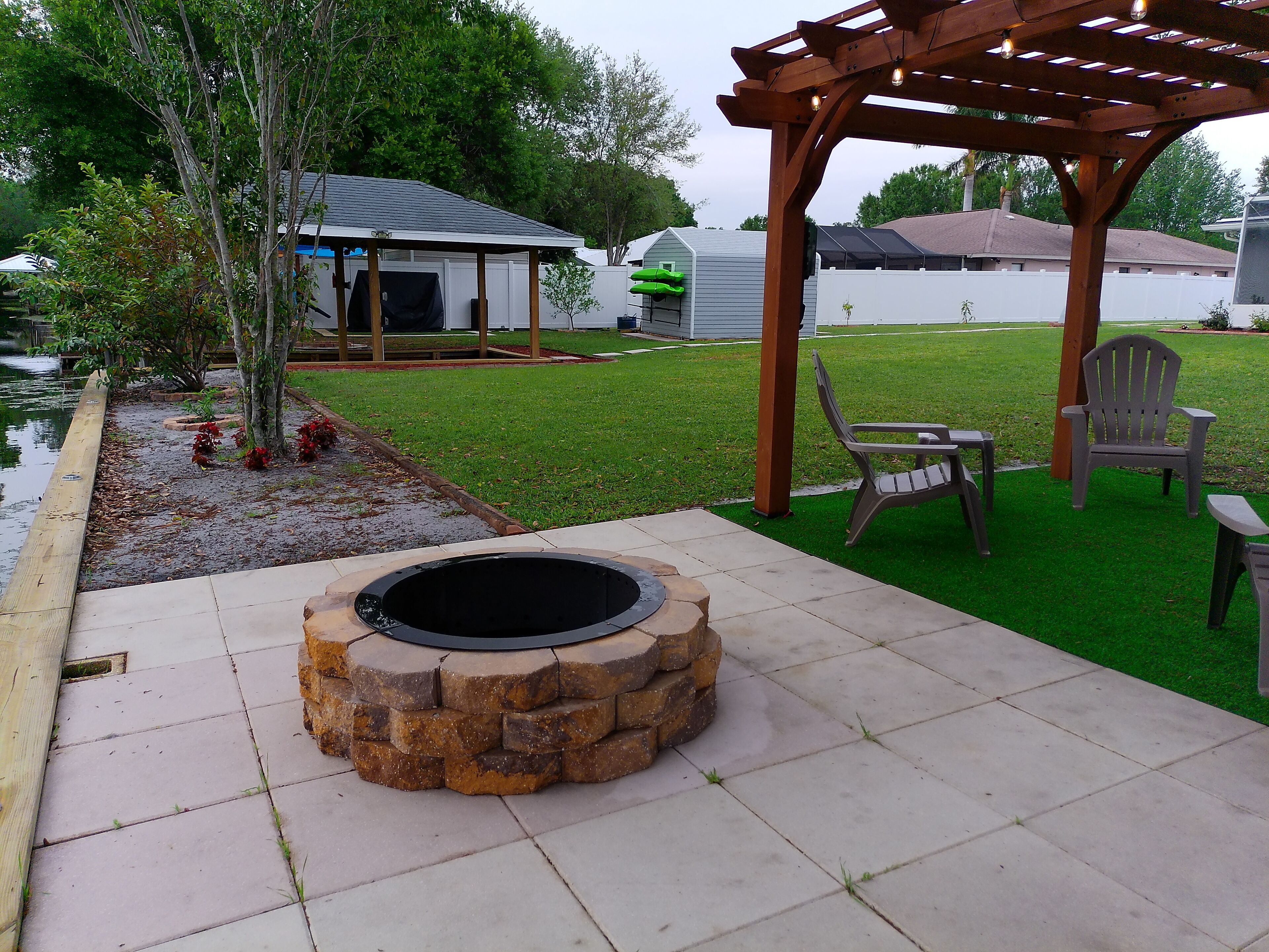 Un pozo de fuego de piedra en un patio trasero con una pérgola y mobiliario exterior.