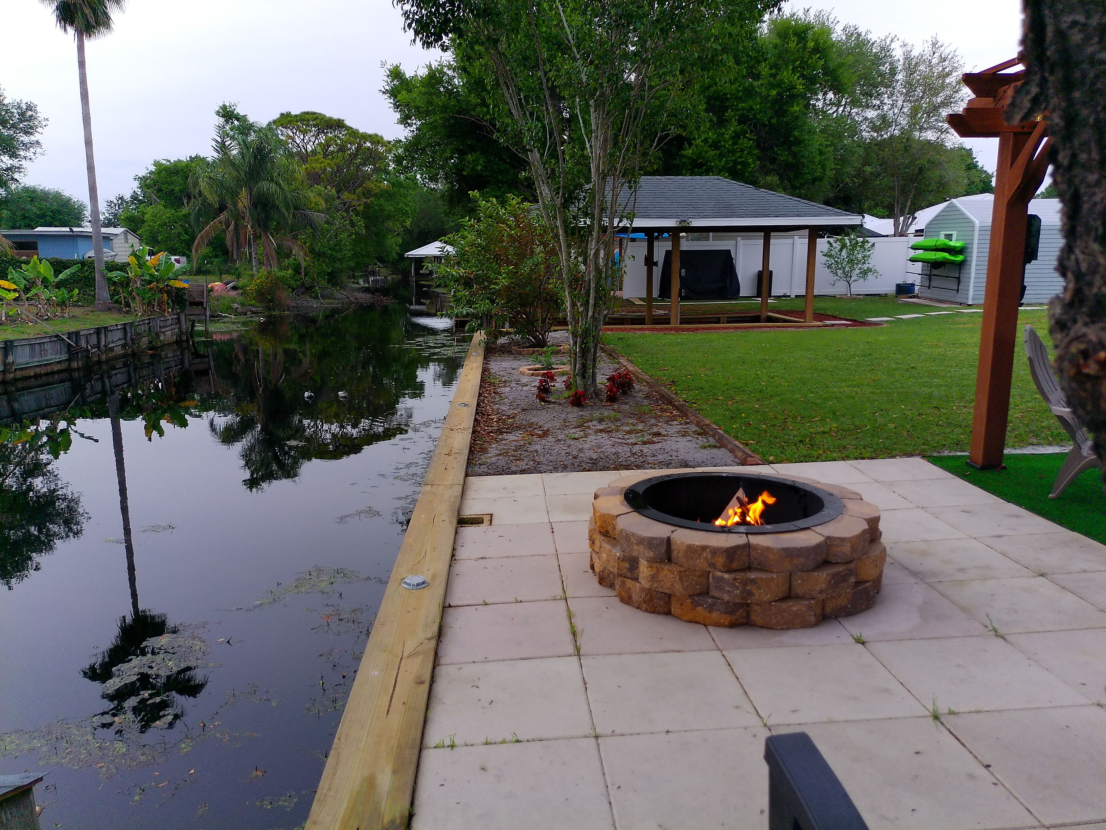 A fire pit in a garden with a pond, surrounded by greenery and a gazebo.
