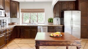 Una cocina con armarios de madera oscura, un refrigerador de acero inoxidable, una mesa de comedor de madera con frutas y una ventana con persianas.