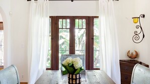 Un comedor con una mesa de madera, cortinas blancas y una ventana con vistas a la vegetación.
