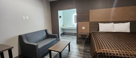 A modern hotel room with a grey sofa, a wooden coffee table, a patterned bedspread, and a built-in headboard.