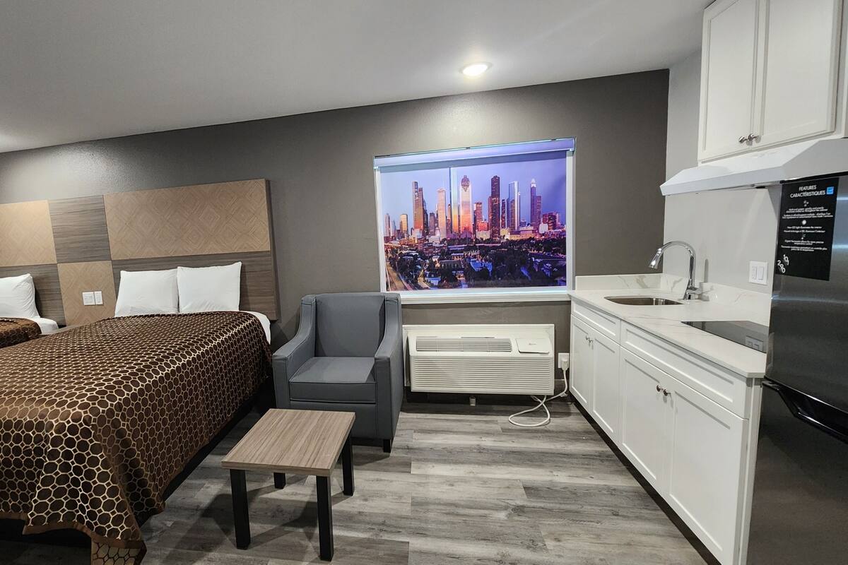 Ein modernes Hotelzimmer mit einem großen Bett, einem grauen Sessel, einem kleinen Tisch, einer Küchenzeile und Blick auf die Stadt.