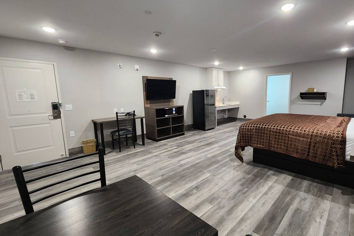 Ein modernes Hotelzimmer mit Bett, Schreibtisch, Stuhl, Fernseher, Kühlschrank und einer kleinen Küchenzeile.