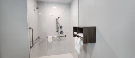 Un espace douche moderne doté d’une porte en verre, d’un pommeau de douche à main, d’un porte-serviettes mural et d’une petite étagère en bois où sont posées des serviettes.